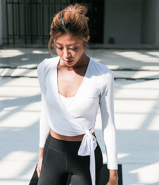 A woman with tied-back hair is looking down while dressed in the white Yoga Comfy Sweater over a white sports bra and high-waisted black leggings. She is standing in a bright, sunlit indoor area with shadows on the floor behind her.