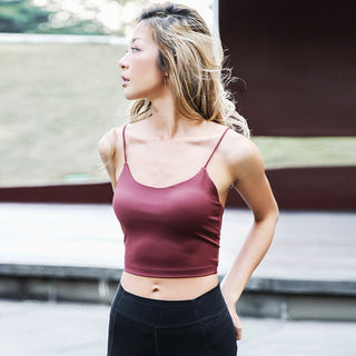 A woman with long, light-colored hair stands outdoors, looking to her left. She wears a maroon Camisole Sports Top and black leggings against a blurred background of grass and a dark brown structure, suggesting it's a bright day.