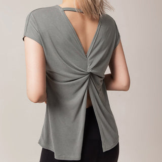 A person wearing a backless green Love Street Style Top is shown from behind. The top features a knotted design at the lower back, with fabric draping down and opening to reveal the upper and mid-back. The person has shoulder-length blonde hair and is also wearing black pants. The background is plain and neutral.