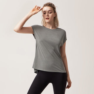 A woman with light-colored hair stands against a plain background, wearing the Love Street Style Top, a loose-fitting, short-sleeve gray garment. She pairs it with black leggings. One of her arms is bent at the elbow, touching above her eyebrow, while the other arm rests at her side. Her pose exudes a casual and relaxed vibe.