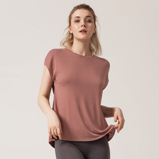 A person with blonde hair is wearing the Love Street Style Top in dusty pink short-sleeve, paired with dark gray leggings. They are looking towards the camera with a neutral expression, holding the hem of the top with one hand while the other hand is slightly raised against a plain, light-colored background.